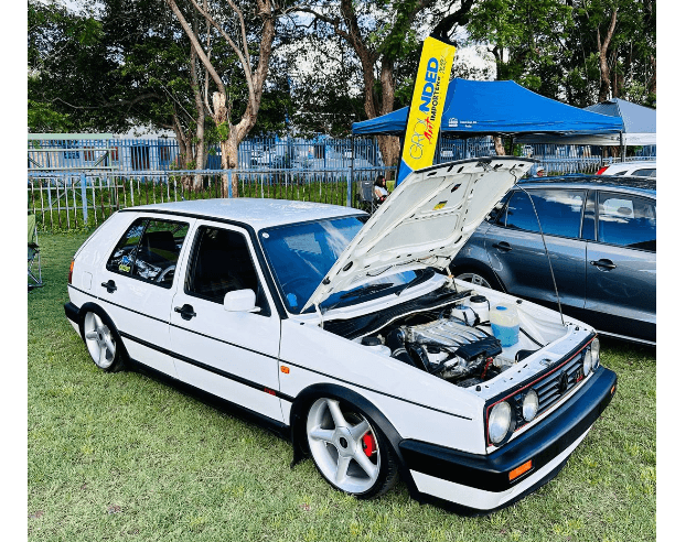 White Volkswagen Golf with its bonnet open, displayed on grass beside other vehicles